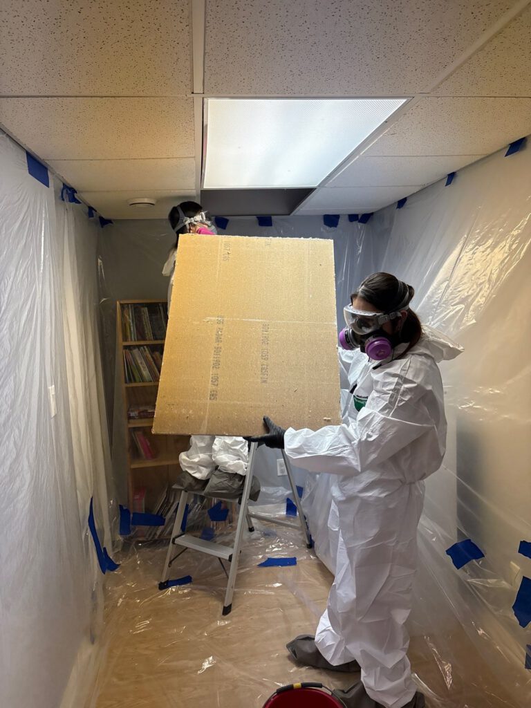 Workers in protective suits carrying a sheet of drywall inside plastic containment