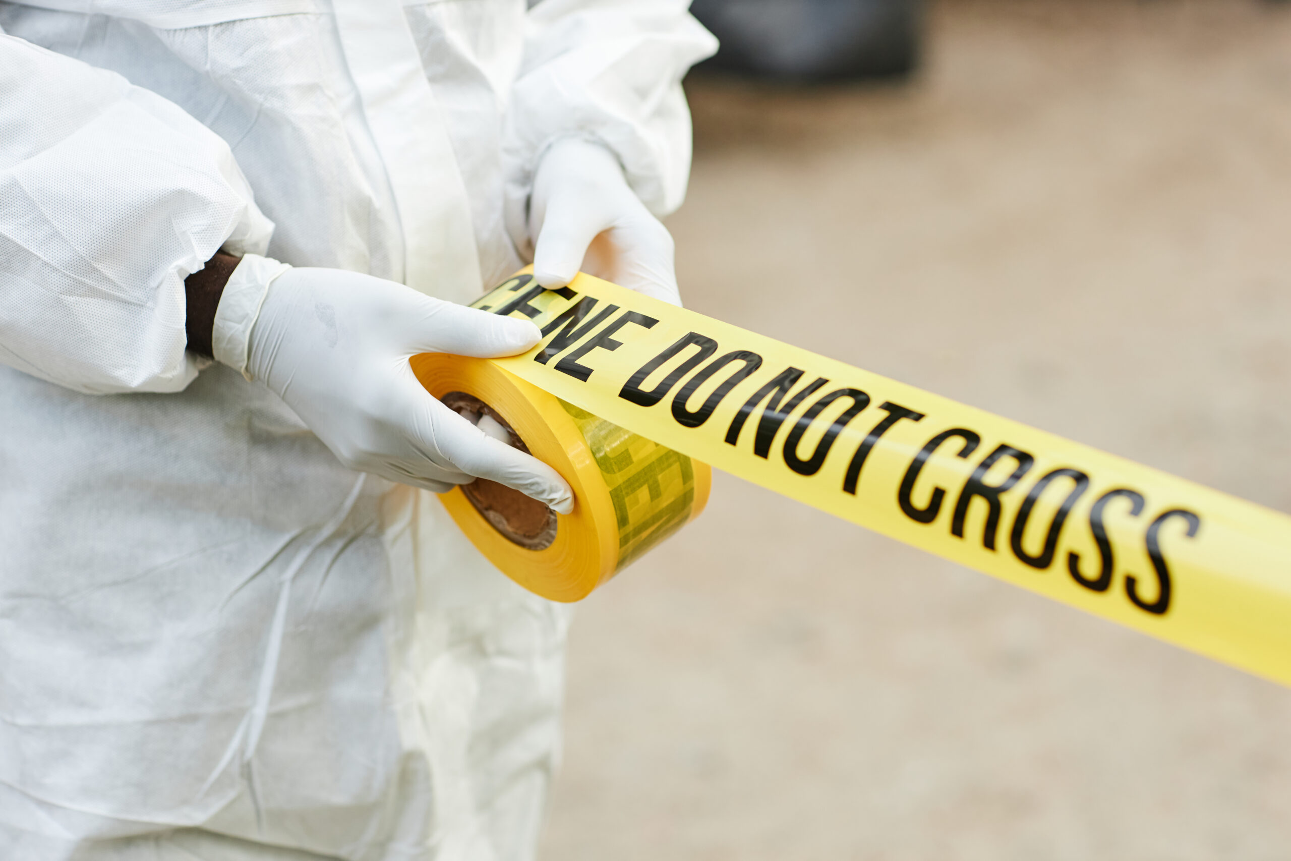 Gloved hands unrolling yellow tape that says Crime Scene Do Not Cross