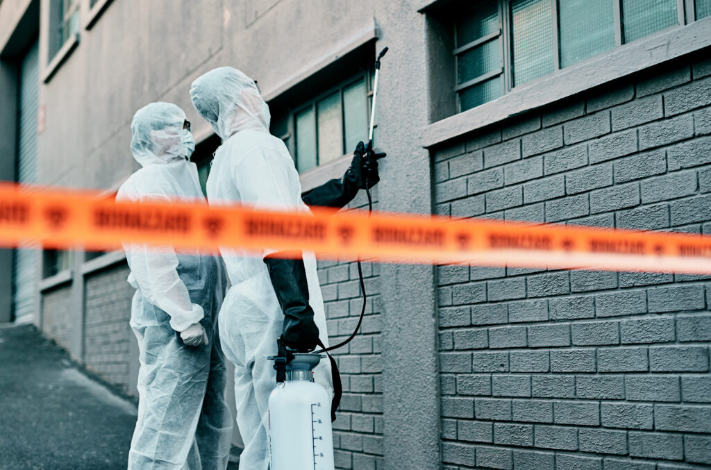 Two workers in protective suits holding a sprayer beside caution tape