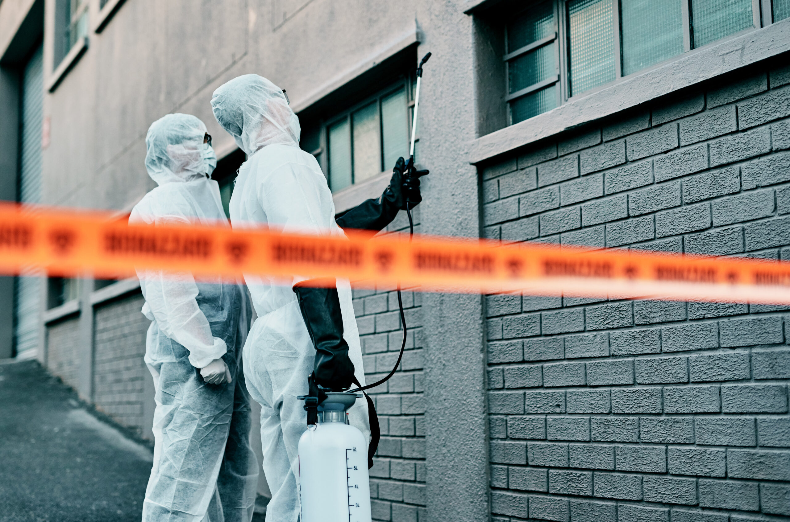 Two workers in protective suits holding a sprayer beside caution tape